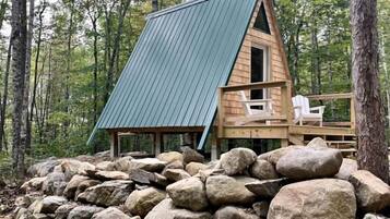 Una cabaña de techo inclinado, rodeada de grandes rocas, y una terraza con sillas.