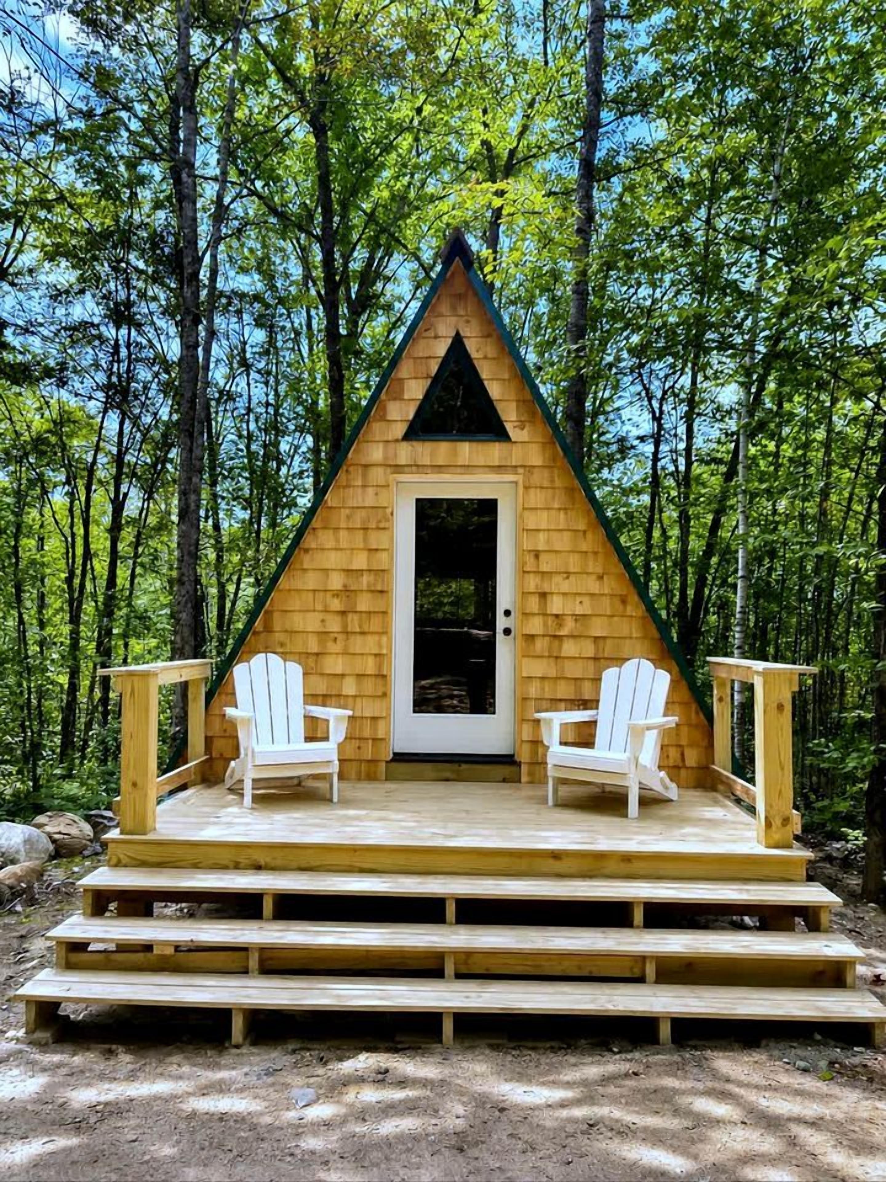 Une cabane en bois avec un toit triangulaire, une porte d’entrée et deux chaises Adirondack blanches sur une terrasse en bois.