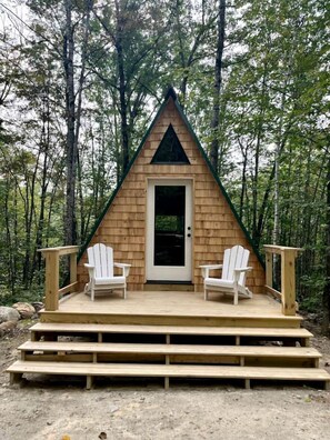 Une cabane en bois avec un toit triangulaire, une porte d’entrée et deux chaises Adirondack blanches sur une terrasse en bois.
