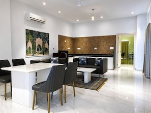 Un salon moderne avec un sol en marbre, un coin repas avec une table et des chaises blanches, un canapé noir agrémenté de coussins à motifs floraux et un grand tableau accroché au mur.