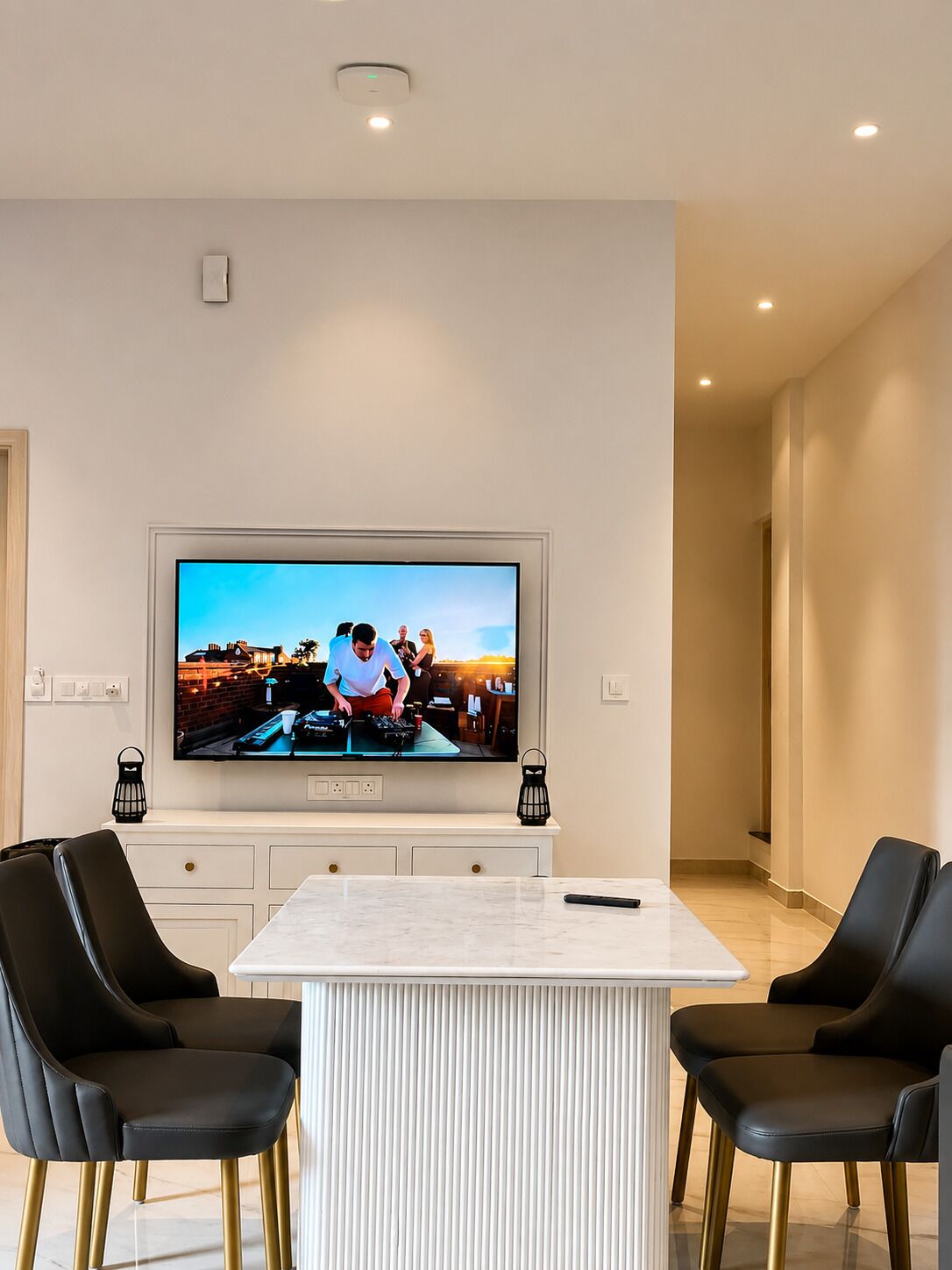 A modern living room with a large flat-screen TV mounted on the wall, a marble-top dining table, and black leather chairs.