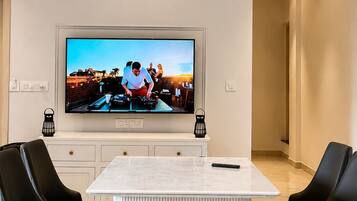 A modern living room with a large flat-screen TV mounted on the wall, a marble-top dining table, and black leather chairs.