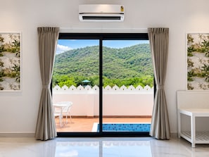 Ein Zimmer mit einer Schiebetür zu einem Balkon mit Pool, weißen Möbeln und Blick auf einen grünen Hügel.