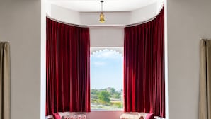 A room with a curved window, red curtains, a cushioned bench, and a view of greenery outside. - Mijmaan Luxury Villa Udaipur (Udaipur)