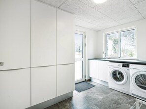Una lavandería con lavadora, secadora, una ventana y una puerta que da a otra habitación.