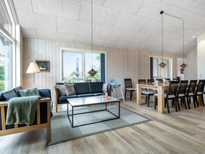 Amplia sala de estar con sofá, mesa de centro y mesa de comedor, todo frente a una pared de madera con ventanas grandes.