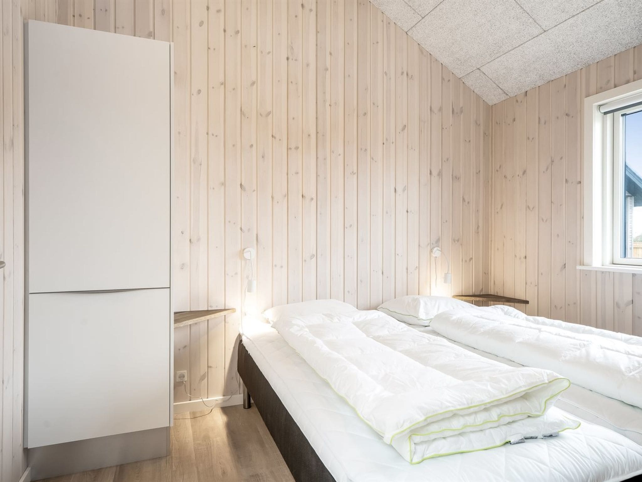 Un dormitorio con dos camas, paredes de madera, un armario, lámparas de noche y una ventana.