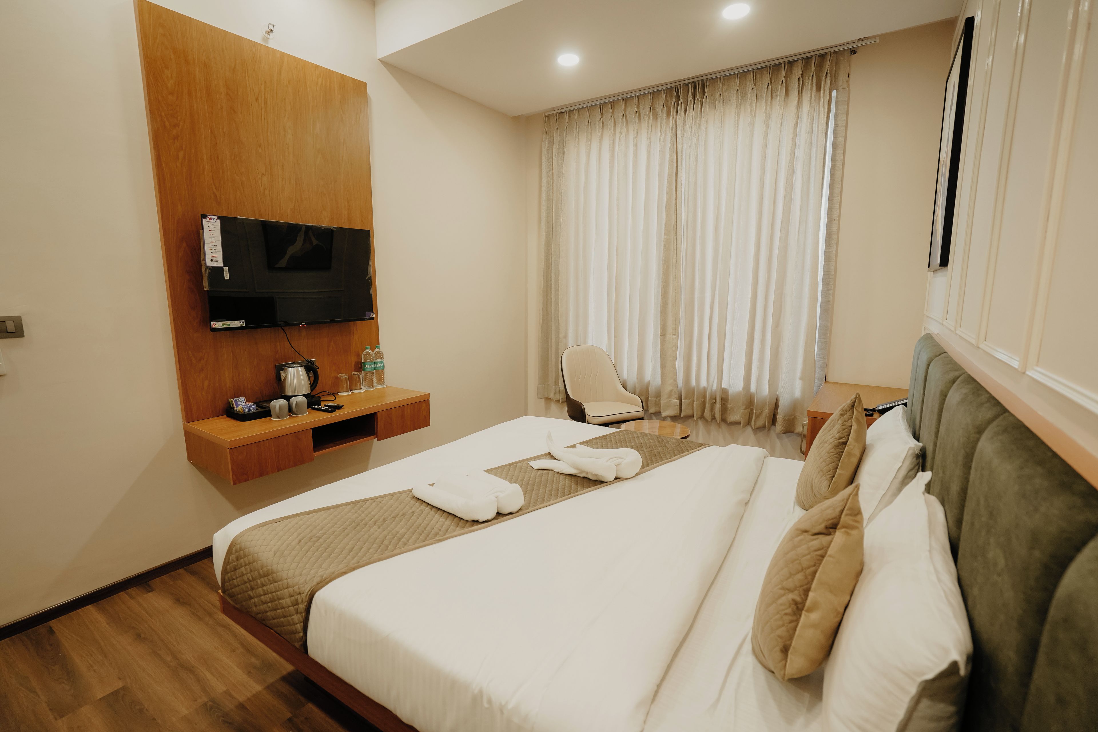 Habitación de hotel con cama, televisor montado en un panel de madera, una silla y una ventana con cortinas.