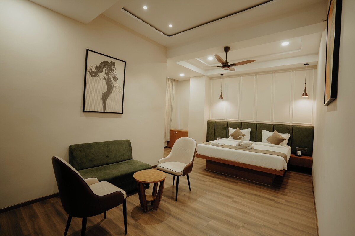 Habitación de hotel con sofá verde, una mesita redonda pequeña, una silla blanca, una cama con cabecera de madera y un ventilador de techo.