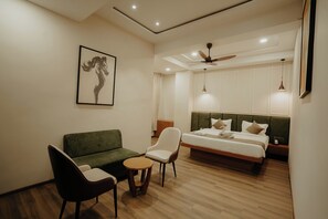 Habitación de hotel con sofá verde, una mesita redonda pequeña, una silla blanca, una cama con cabecera de madera y un ventilador de techo.