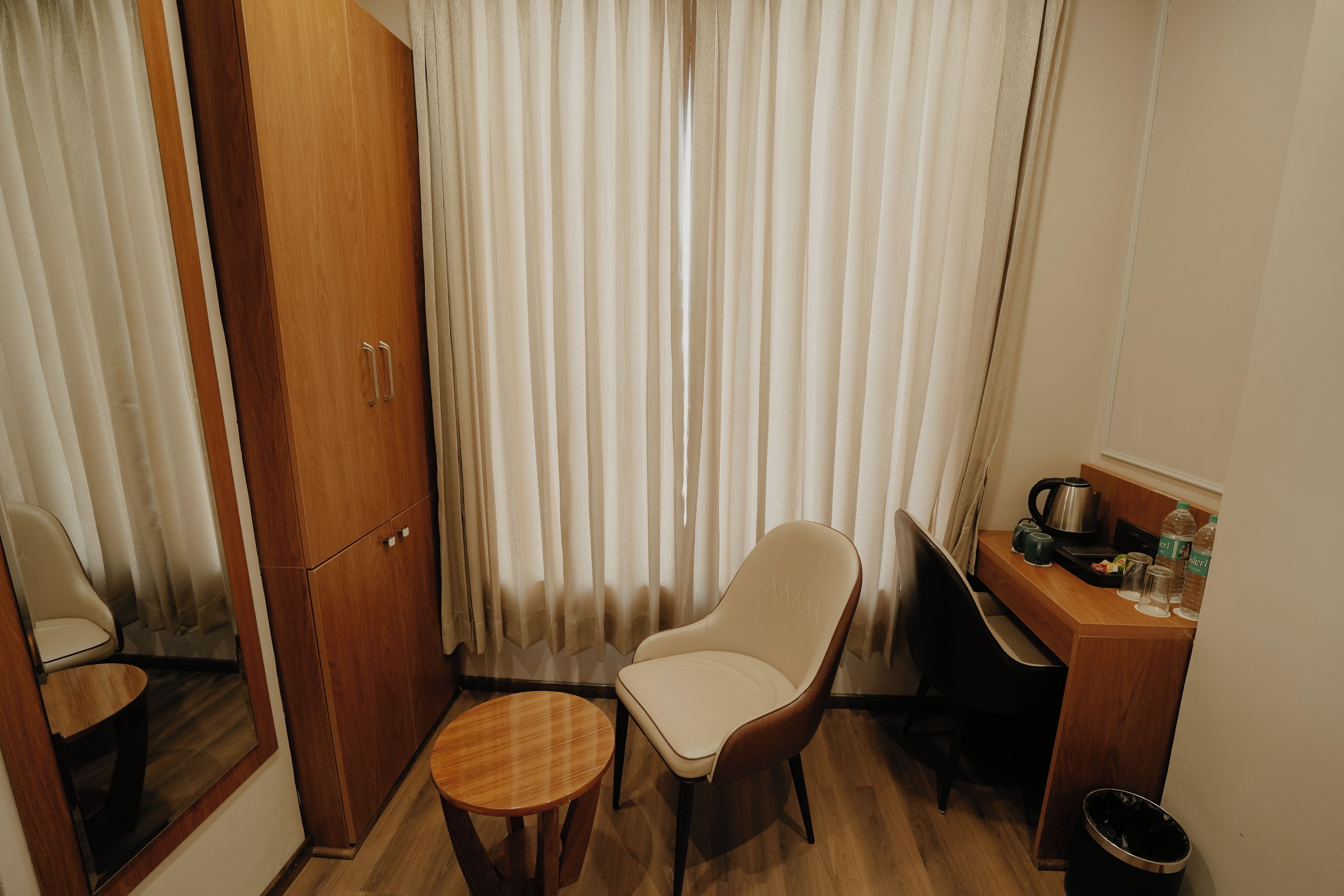 Una habitación pequeña con un armario de madera, una silla, una mesita redonda, un escritorio con una tetera, tazas y botellas.