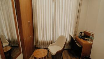Una habitación pequeña con un armario de madera, una silla, una mesita redonda, un escritorio con una tetera, tazas y botellas.