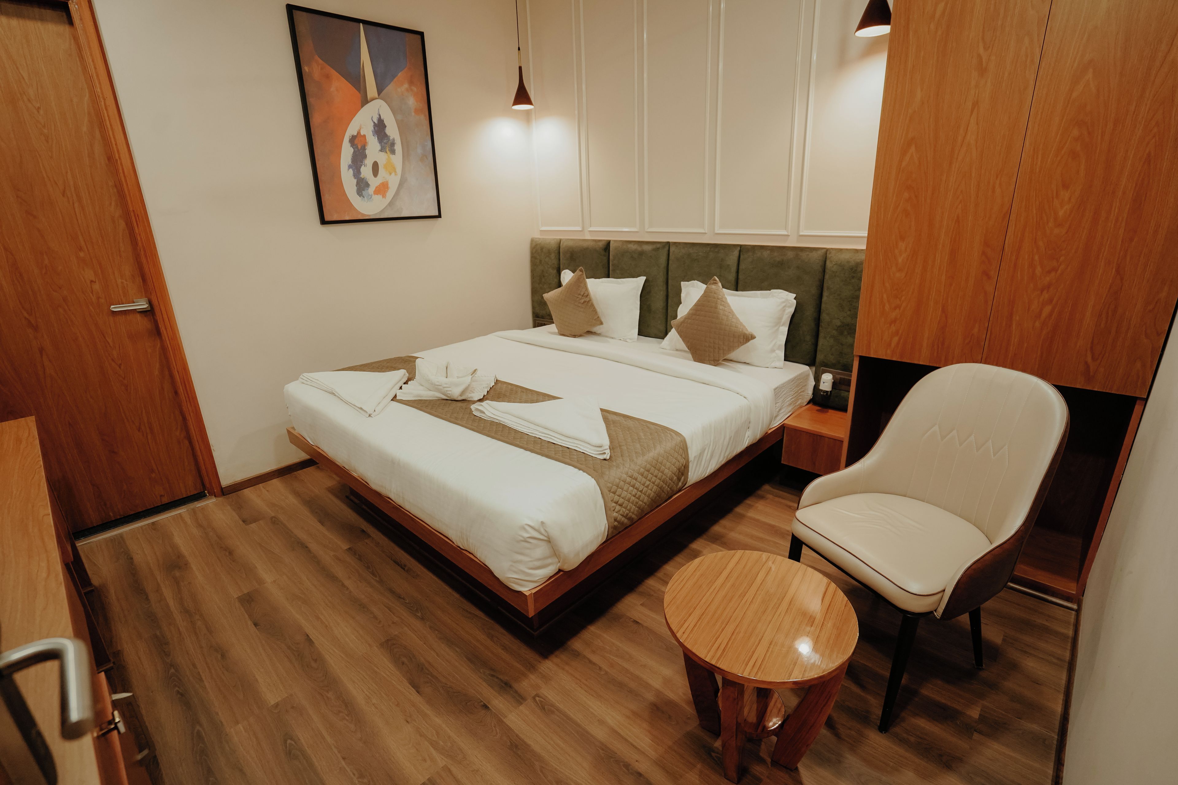 A neatly made bed with pillows and a blanket, a wooden chair, a small round table, and a painting on the wall.