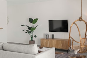 Ein modernes Wohnzimmer mit einem grauen Sofa, einer Holzkabine, einer großen Topfpflanze, einem hängenden Korbstuhl und einem an der Wand montierten Flachbildfernseher.