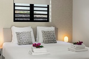 A neatly made bed with white linens, two patterned pillows, and a stack of folded towels. - Apartment Ayana Mambo Beach (Willemstad)