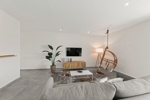 En moderne stue med en grå sofa, et hvidt sofabord, et træskab, en stor stueplante og en gulvlampe.
