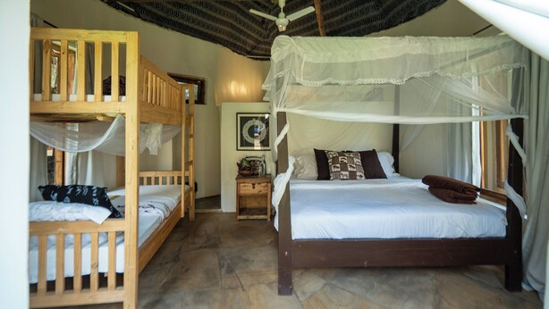 Una habitación con una cama nido de madera, una cama individual con dosel, una mesita de noche pequeña y un estante fijado a la pared.