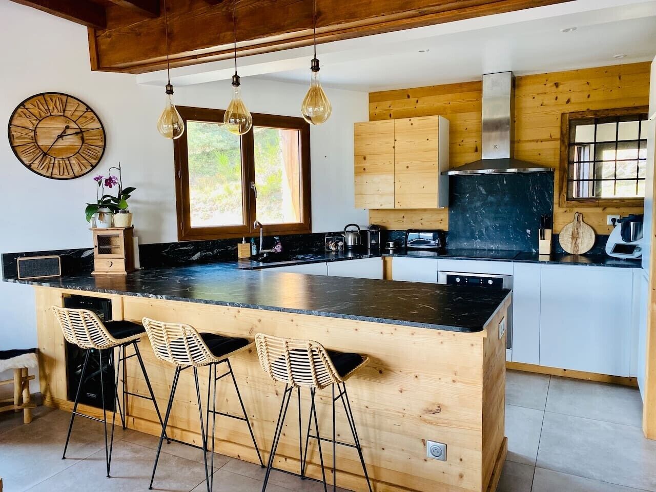 Une cuisine moderne dotée d’un îlot central, de tabourets de bar, de meubles de rangement en bois et de luminaires suspendus.