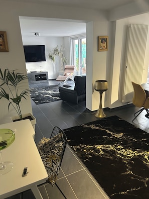 Een moderne woonkamer met een eethoek, een aan de muur bevestigde televisie en een groot raam dat zorgt voor veel daglicht.
