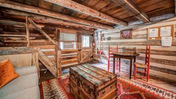A rustic log cabin interior with a staircase, a wooden chest, a dining table with chairs, and a sofa.
