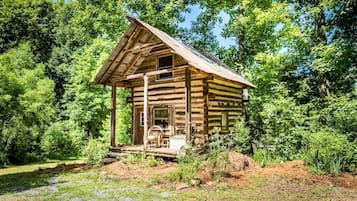 A rustic log cabin with a porch and a chimney, surrounded by dense forest.