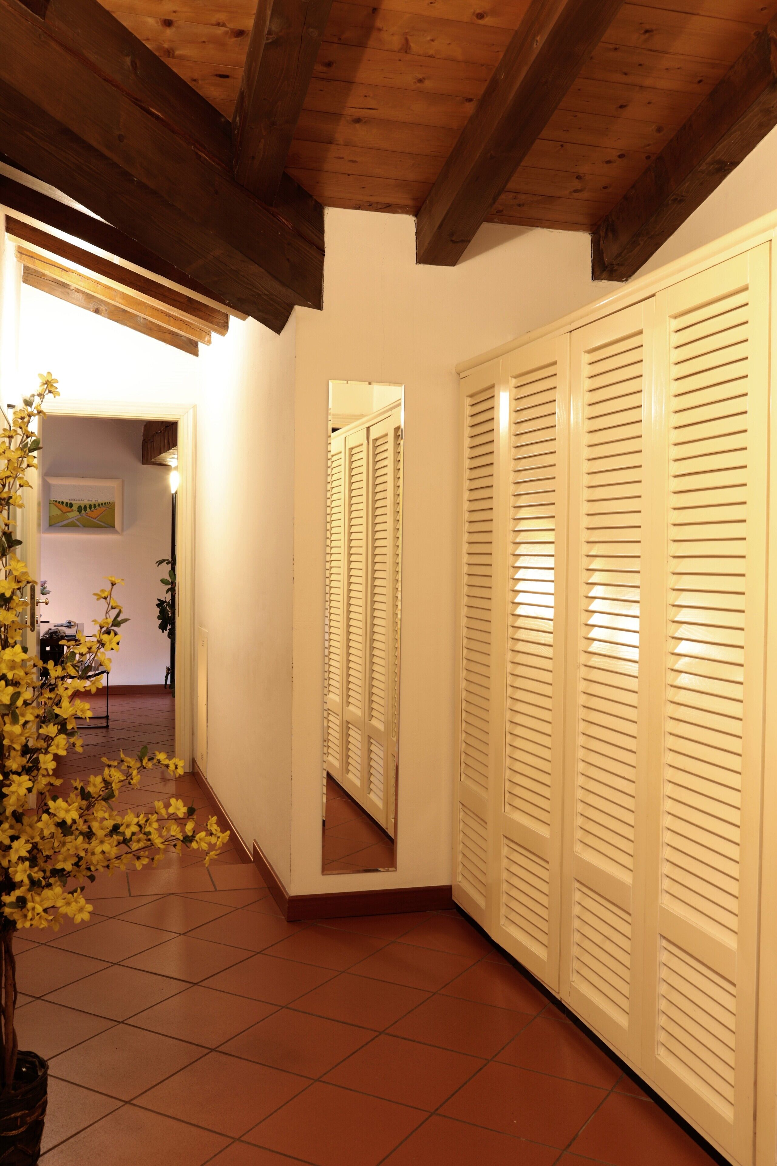 Un couloir avec un plafond en bois, des murs blancs et un sol carrelé. On y trouve une plante en pot avec des fleurs jaunes, un miroir qui reflète un placard aux portes à persiennes, et une porte qui donne sur une autre pièce.