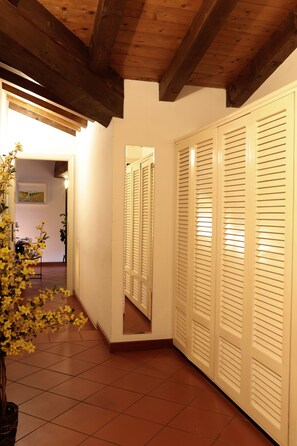 A hallway with a wooden ceiling, white walls, and a tiled floor. There is a potted plant with yellow flowers, a mirror reflecting a closet with louvered doors, and a door leading to another room. - The Rose Apartment - Bologna (Bologna)