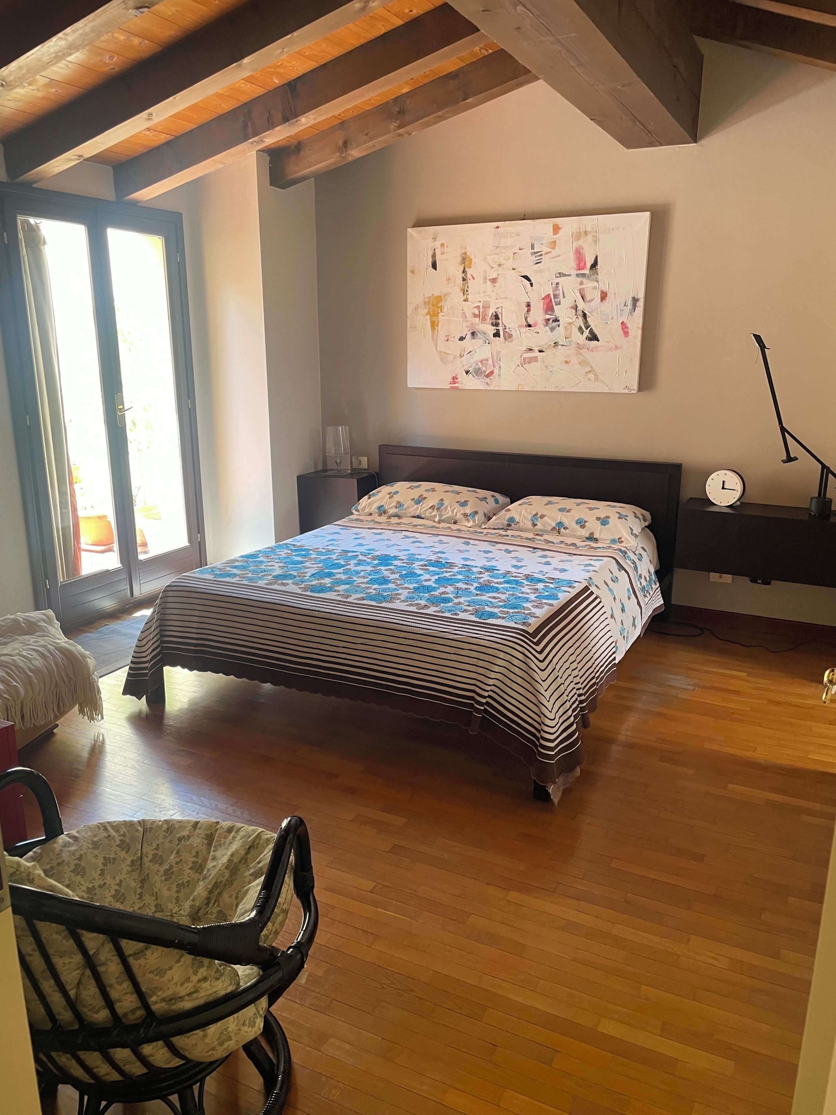 A bedroom with a bed, a chair, a nightstand, a wall clock, and a large abstract painting.