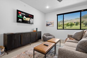 A living room with a large TV displaying streaming services, a dark wooden cabinet, a gray bean bag, a beige sofa, and a window with a mountain view. - Greenside Retreat Pt C207|Ski &Summer Getaway (Eden)