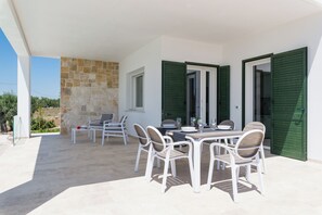 A covered outdoor dining area with white furniture and green shutters, stone wall, and a view of greenery. - Charming Villa with Pool by Wonderful Italy (Ostuni)