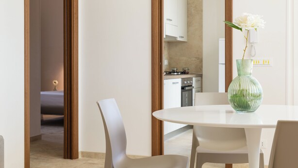 Ein moderner Essbereich mit einem weißen Tisch und Stühlen, einer grünen Vase mit einer weißen Blume sowie einer Küche im Hintergrund.