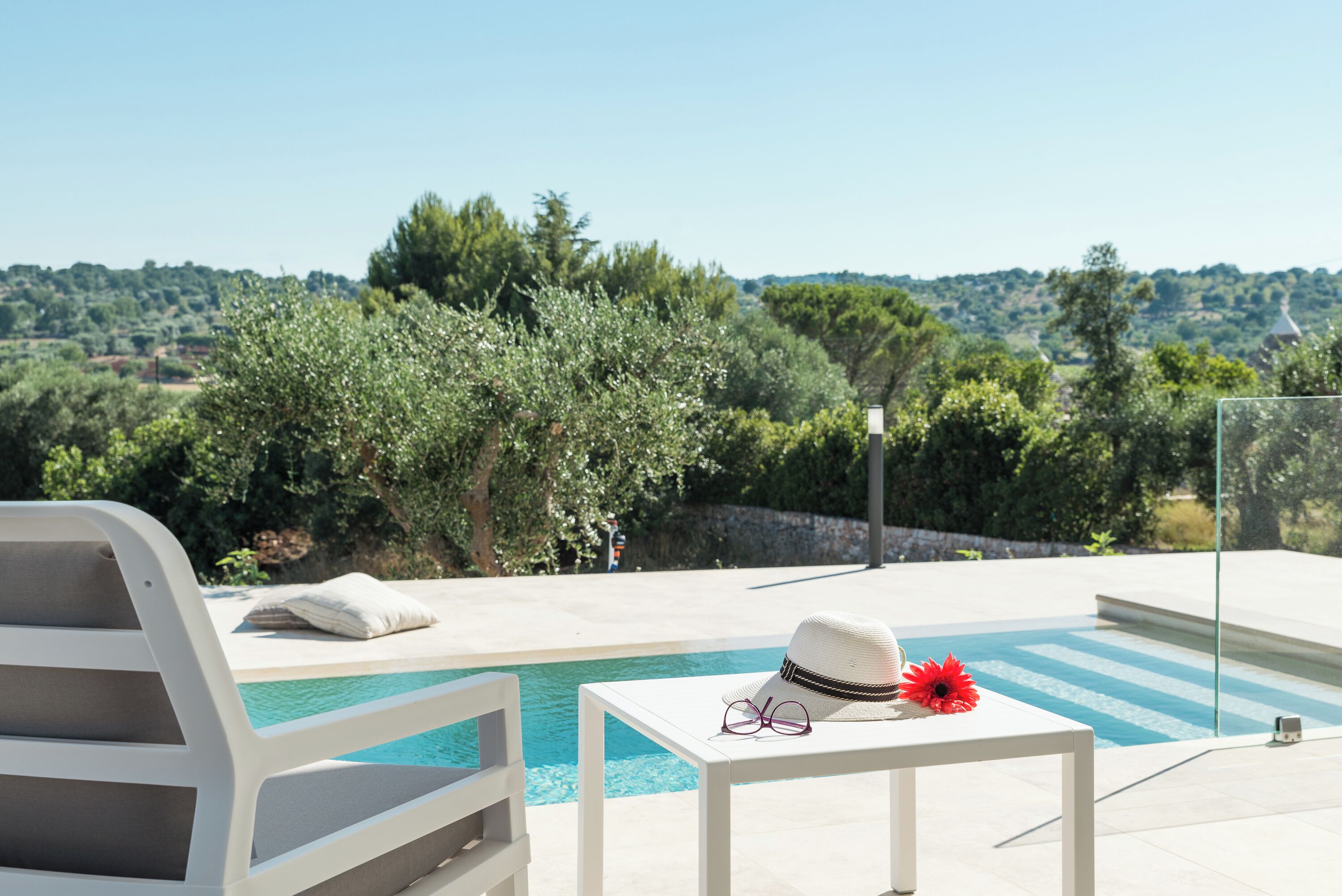 A poolside area with a white lounge chair, a small table with a hat and glasses, and a view of a hilly landscape.