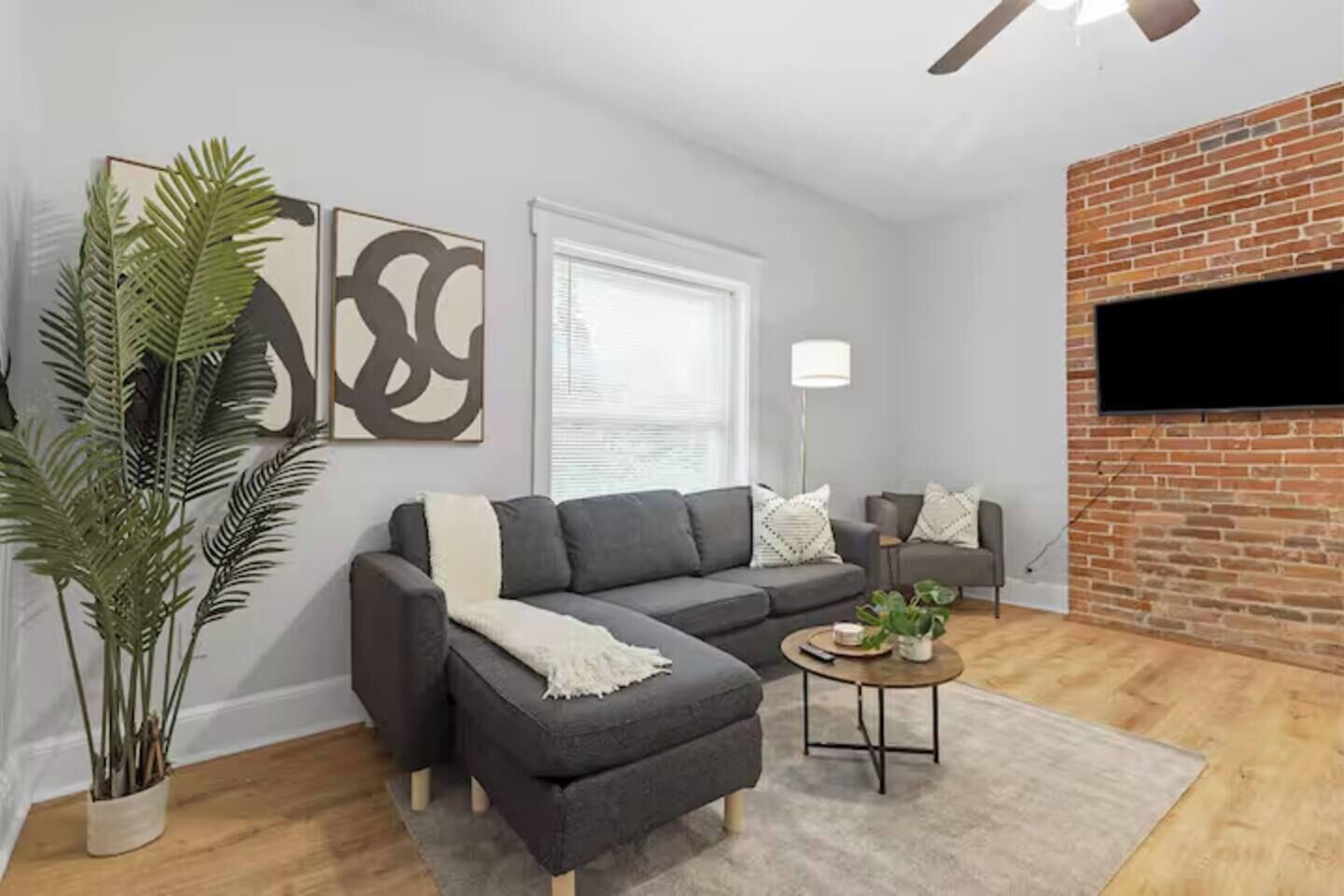 A living room with a grey sofa, a coffee table, a potted plant, and a brick wall.