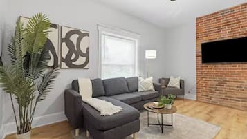 A living room with a grey sofa, a coffee table, a potted plant, and a brick wall.