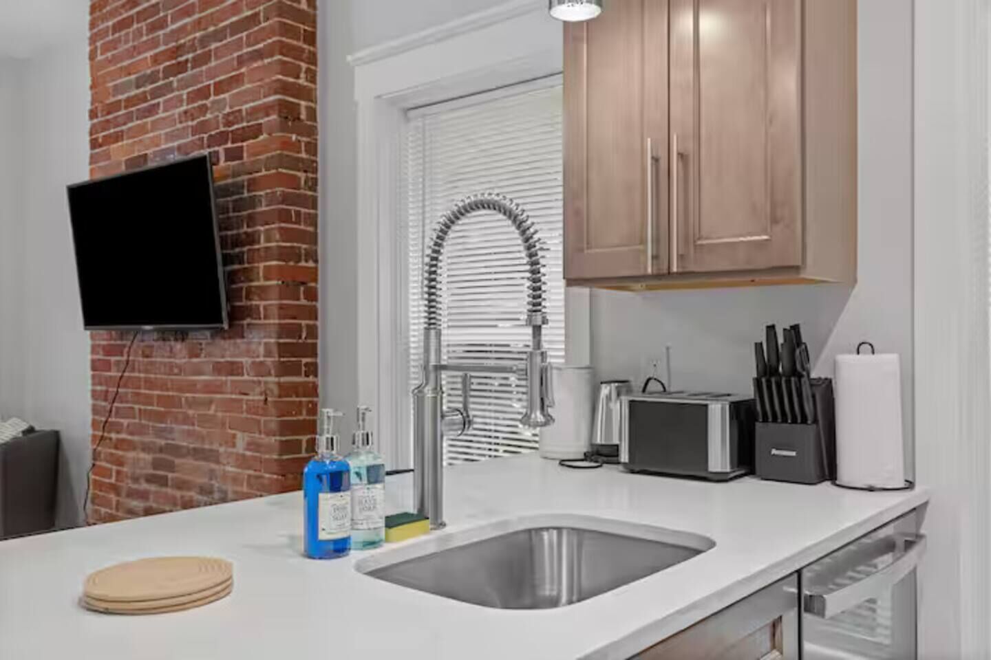A modern kitchen with a brick wall, a flat-screen TV, a stainless steel sink, and wooden cabinets.