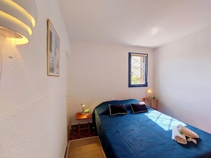 Ett litet, smalt rum med en enkel säng, ett litet bord med en lampa, ett fönster med utsikt och en tavla som hänger på väggen.