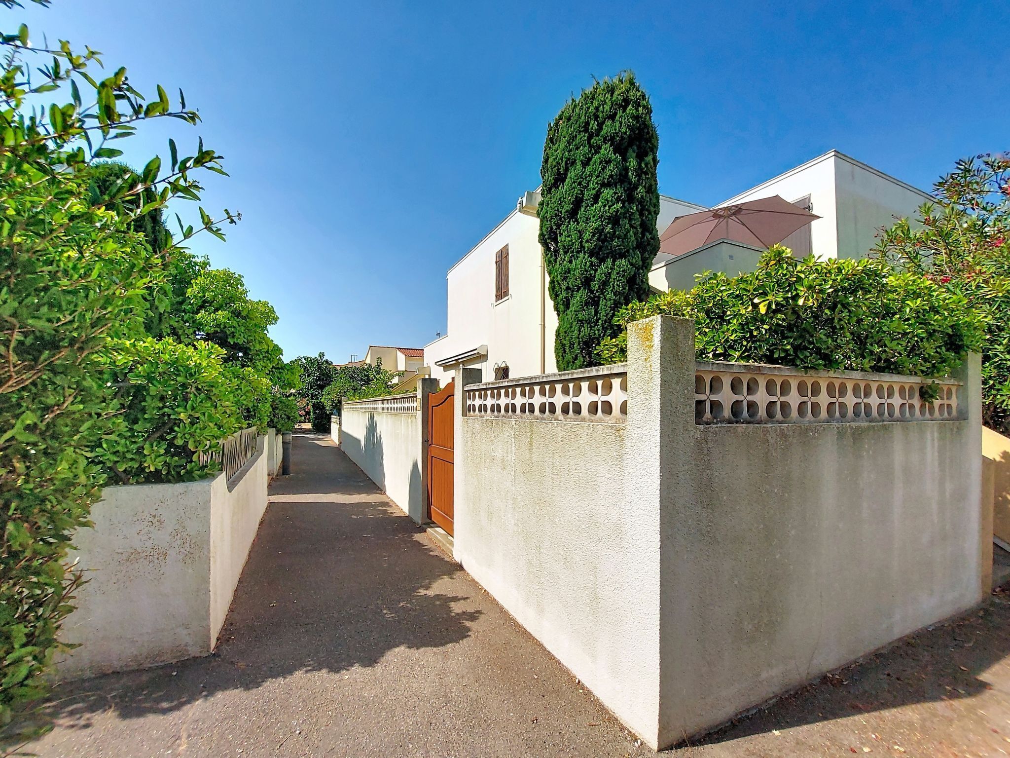 Une rue résidentielle étroite, bordée de maisons des deux côtés, avec un ciel dégagé et un grand arbre au centre.