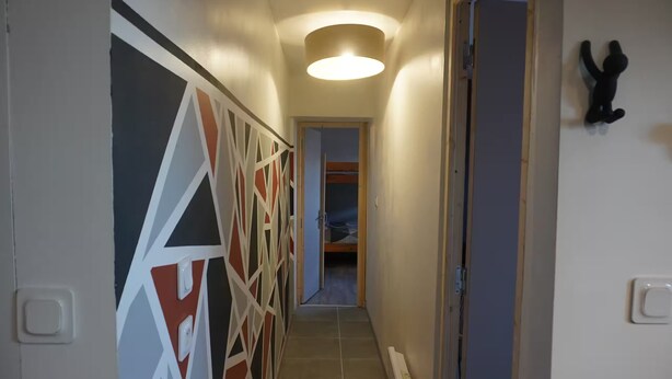 A narrow hallway with a geometric-patterned wall on the left, a door on the right, and a bed visible at the end. - Family-friendly city apartment with 2 free private indoor parking spaces (Beuvrages)