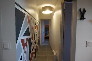 A narrow hallway with a geometric-patterned wall on the left, a door on the right, and a bed visible at the end.