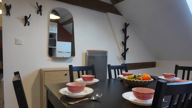 A dining area with a dark wooden table, white plates, and red patterned bowls. There are two chairs, a mirror, and a wall-mounted heater.