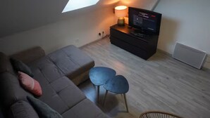 A loft-style room with a high ceiling, a skylight, a sofa, a small table, a TV on a stand, and a radiator. - Family-friendly city apartment with 2 free private indoor parking spaces (Beuvrages)