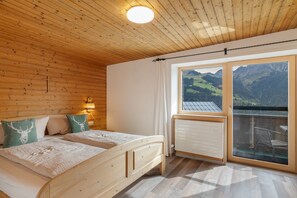 A bedroom with a wooden ceiling, a bed with two pillows, a nightstand with a lamp, and a window with a view of mountains.