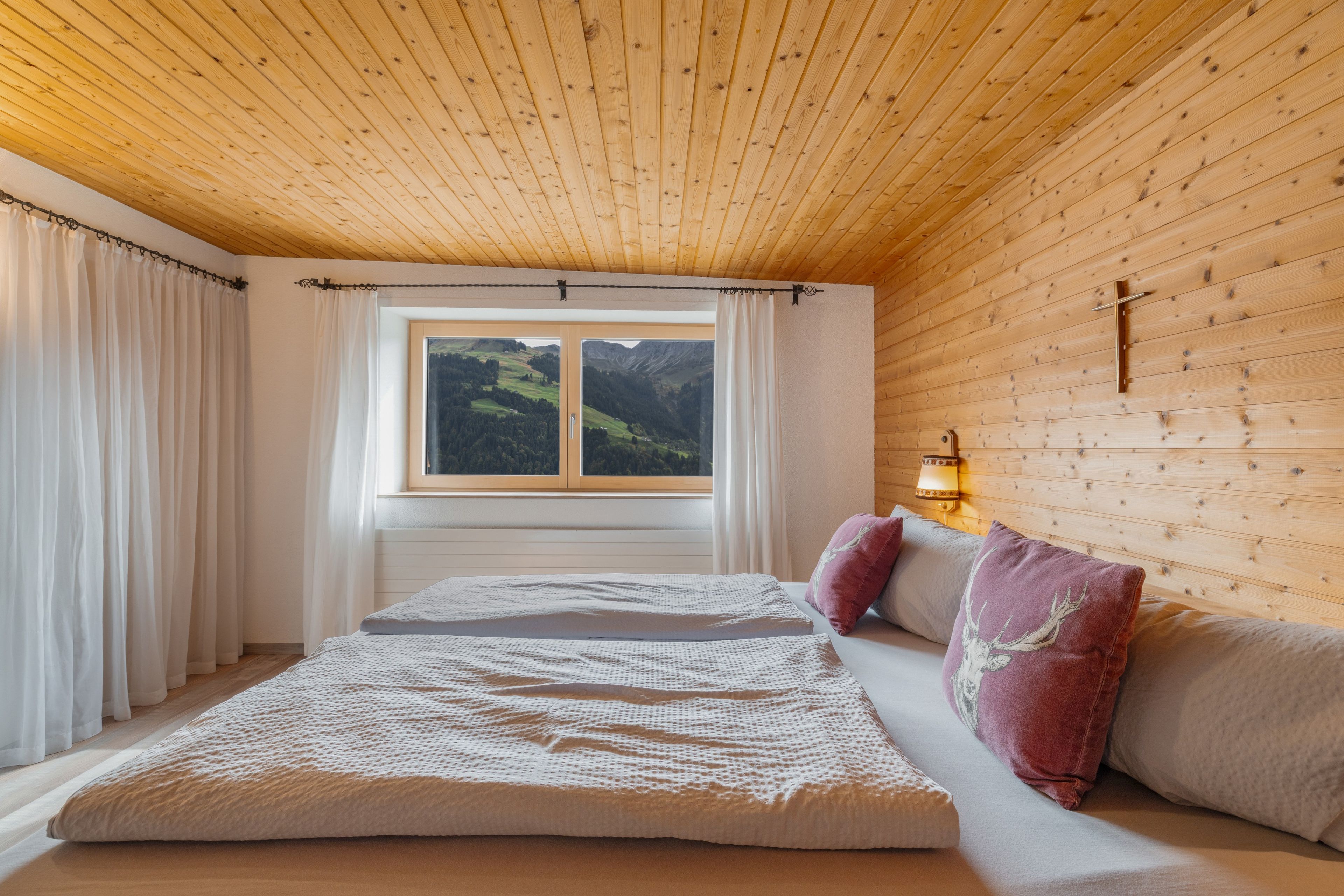Un dormitorio con techo de madera, una cama con colchón blanco, dos almohadas y una ventana con vistas a las montañas.