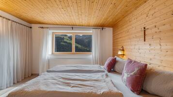 Un dormitorio con techo de madera, una cama con colchón blanco, dos almohadas y una ventana con vistas a las montañas.