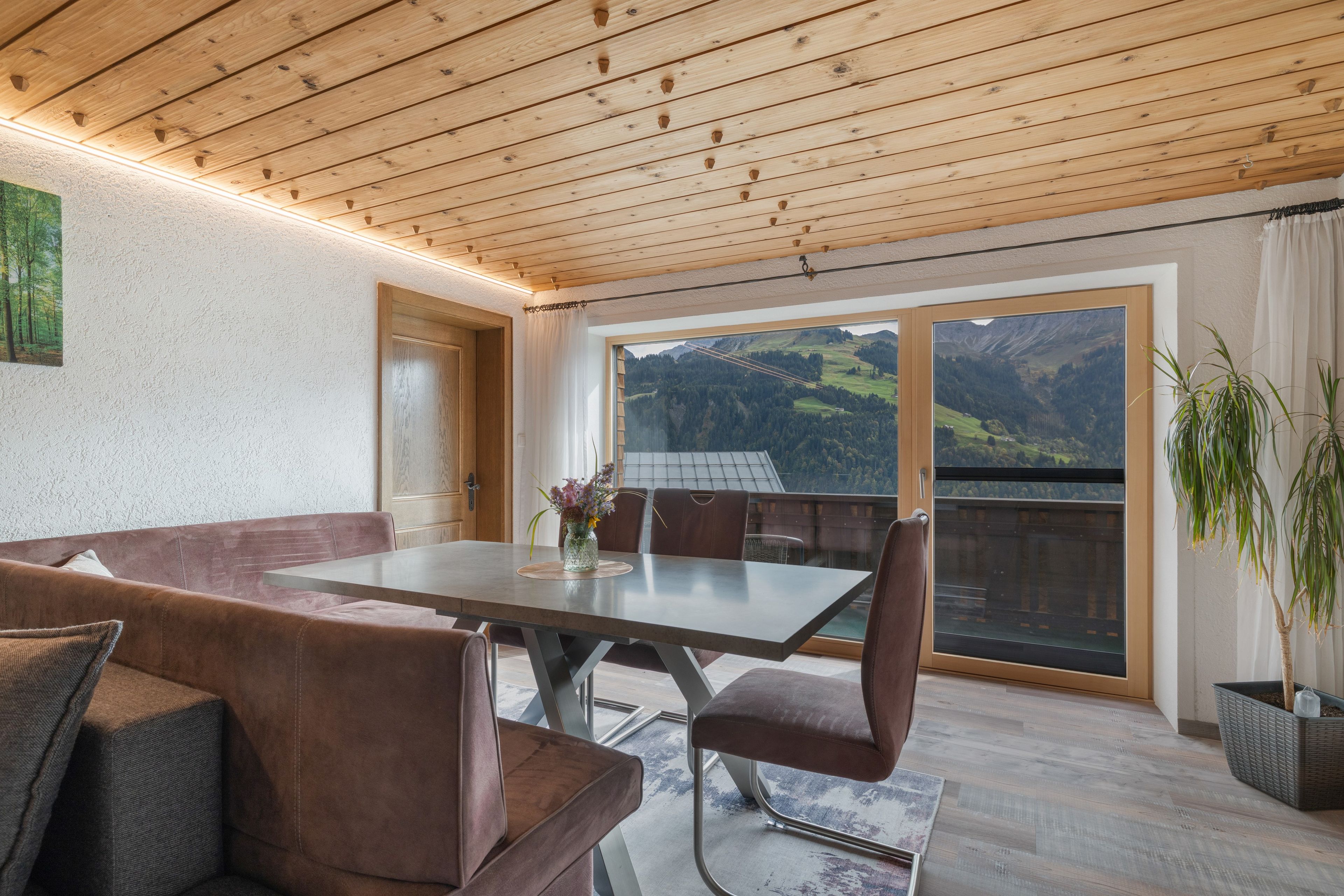 Un espace repas comprenant une table et des chaises, un plafond en bois et une vue sur les montagnes grâce à de grandes fenêtres.
