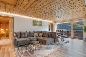 Un soggiorno moderno con soffitto in legno, un divano spazioso, un tavolo da pranzo e vista su un paesaggio montuoso attraverso una grande finestra.