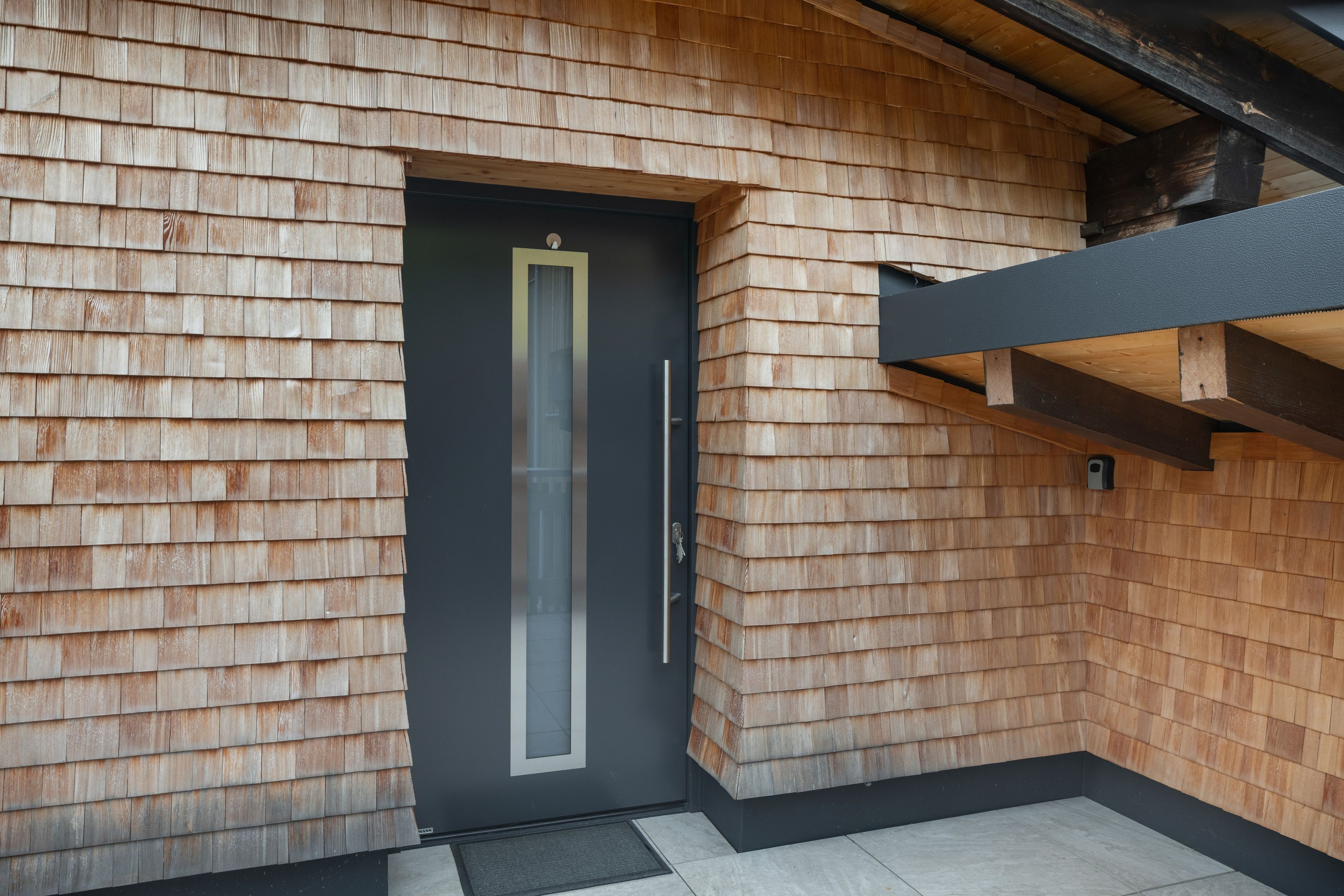 Un bâtiment moderne avec une façade recouverte de bardeaux de bois, une porte de couleur sombre dotée d’une poignée verticale et une structure en poutres métalliques apparentes.