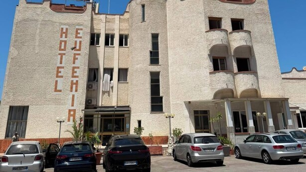 A hotel with a sign reading 'HOTEL' and 'HOTELERA' on the facade, multiple parked cars, and a clear blue sky.