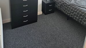Une chambre avec une commode noire, une table de chevet, un lit recouvert d’une couverture à carreaux et un vase rempli de fleurs posé sur la commode.