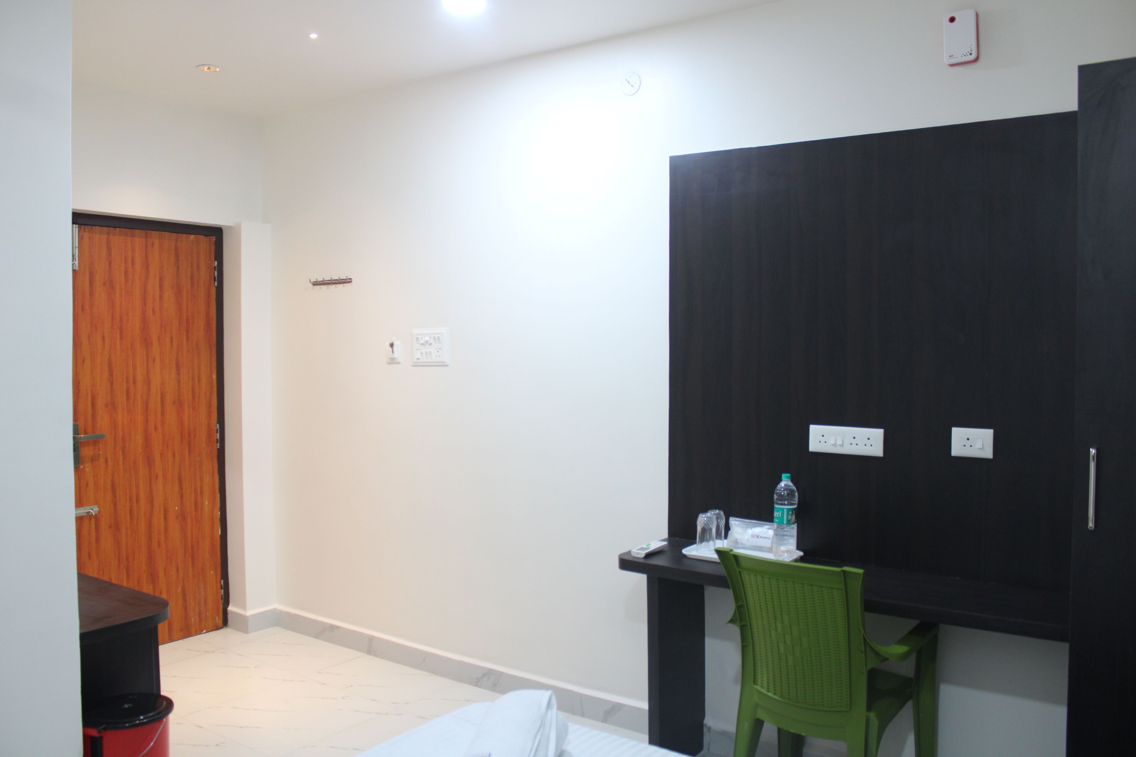 A hotel room with a wooden door, a desk with a chair, a black wardrobe, and a small table with a water bottle.